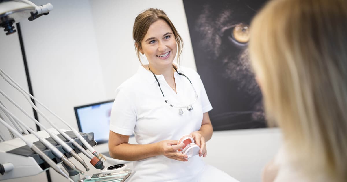 A dentist dressed in white scrubs, holding a model of teeth, talking to a patient. 
