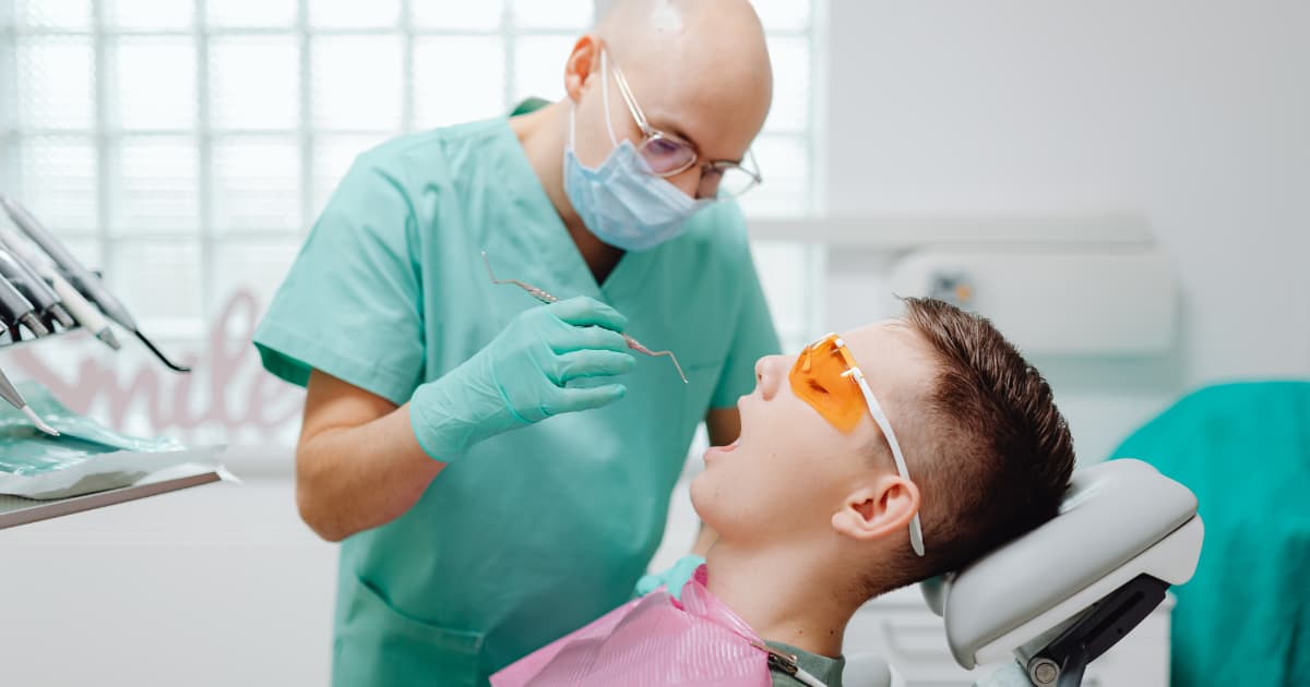 An older teenage boy sits in a dentist practice chair while a dentist looks in his mouth
