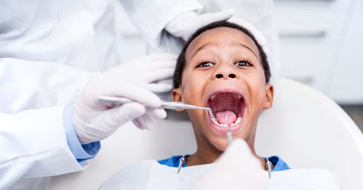 a boy about 10 years old opens wide while he sits in a dentist exam chair and a dentist uses tools for an exam.