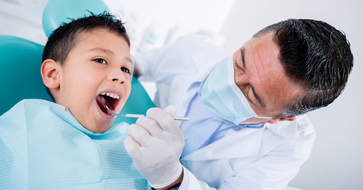 A boy sits in a dental exam chair while a dentist wearing a mask uses a mirror to look at his teeth.