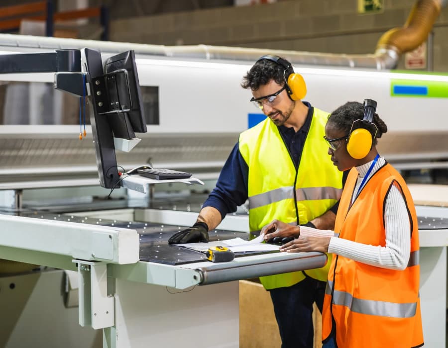 Two manufacturing workers wearing safety glasses, hearing protection, and high-visibility vests review paperwork beside industrial equipment.