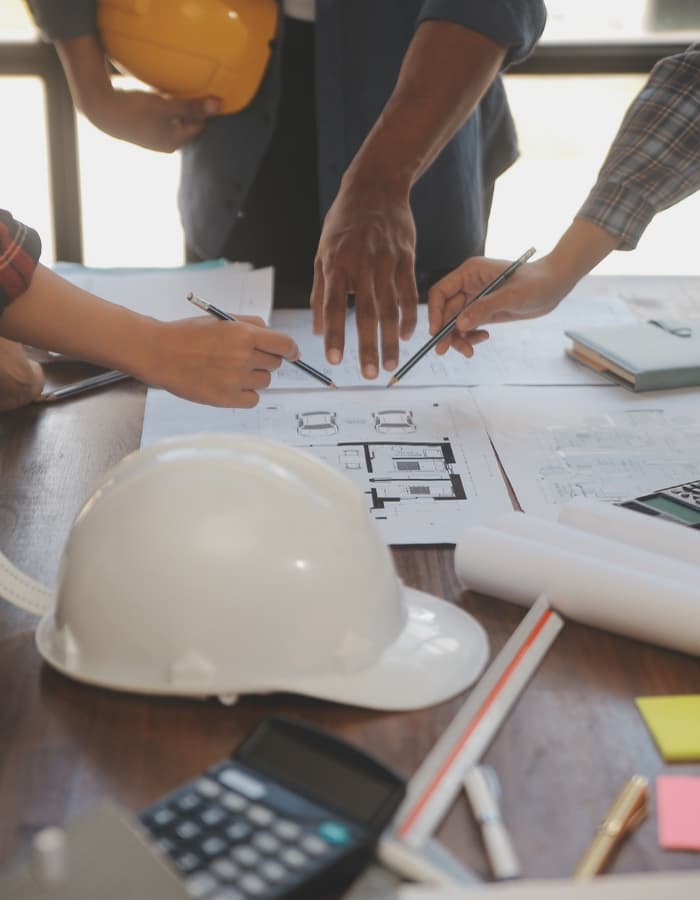 drafting plans on a table with a white hardhat next to it. People are pointing to the draft plans and you can see their hands